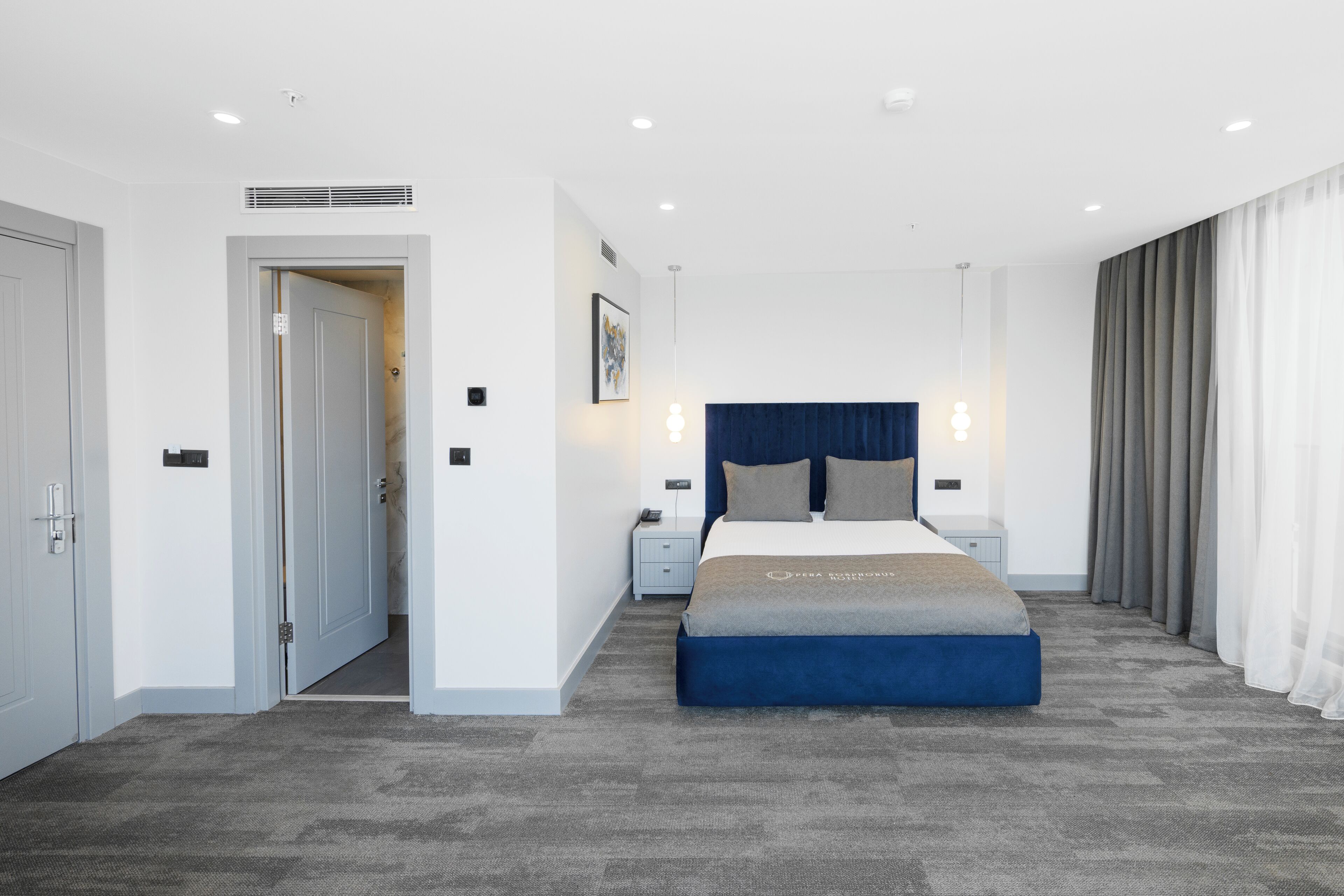 Une chambre d’hôtel moderne avec un grand lit, une table de chevet, une commode, une porte et une fenêtre avec des rideaux.