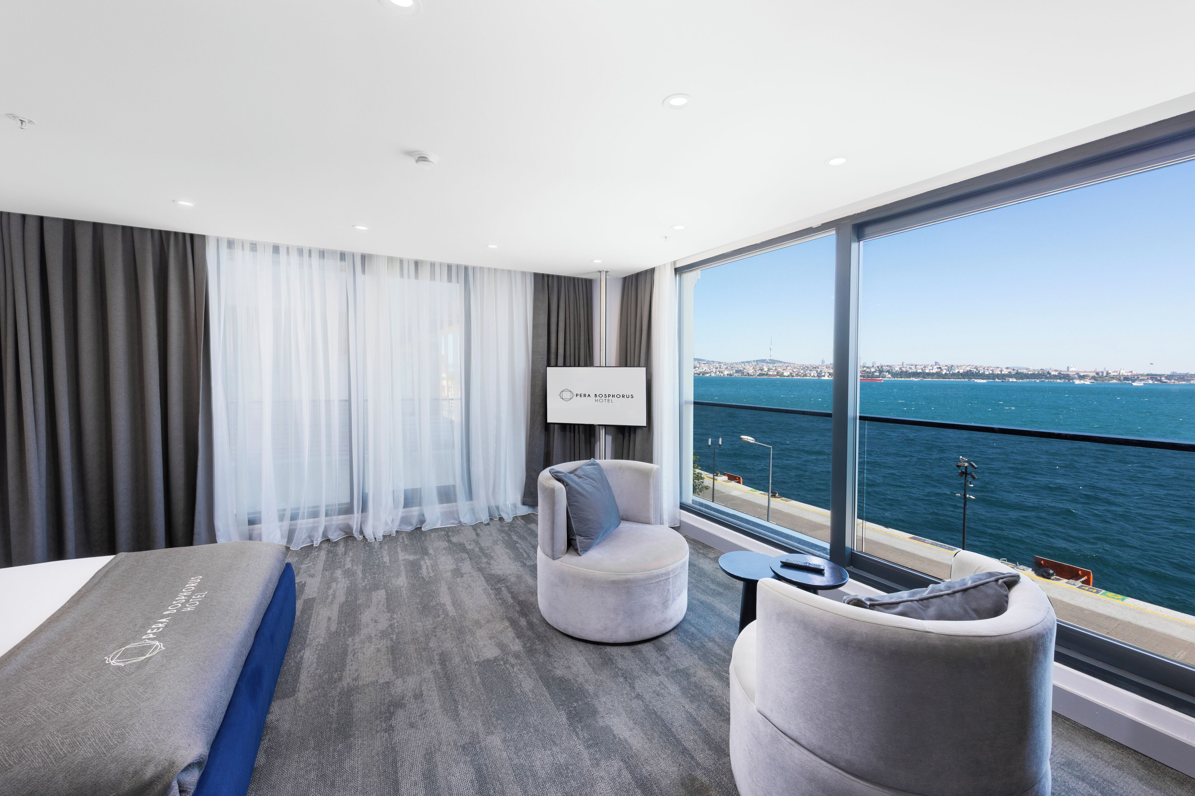 A modern hotel room with a large window offering a view of the sea and city skyline.