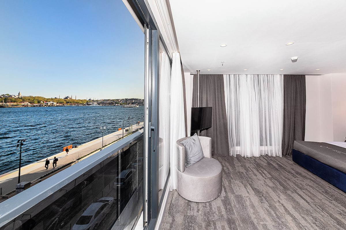 A modern hotel room with a large window overlooking a waterfront, a comfortable seating area, and a flat-screen TV.