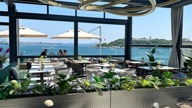 An outdoor seating area with tables and chairs, surrounded by potted plants, overlooking a body of water with a distant view of a cityscape. - Pera Bosphorus Hotel (Istanbul)