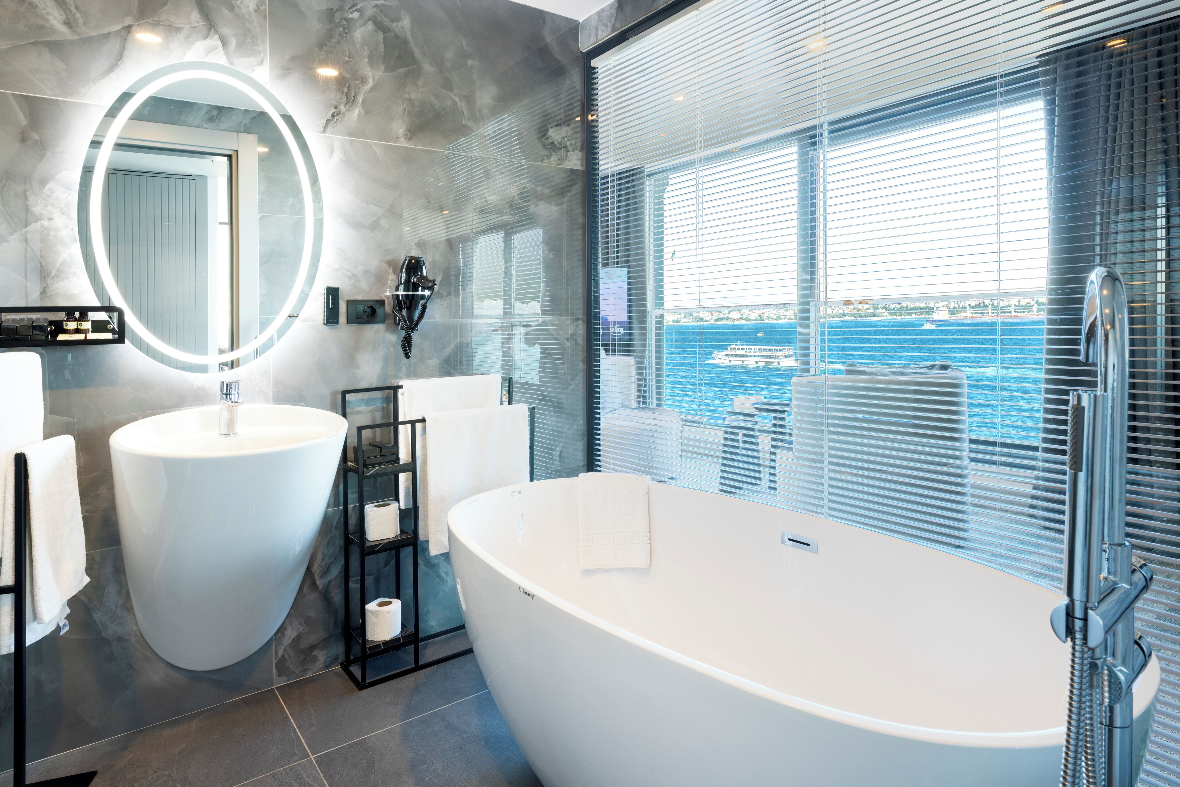 A modern bathroom with a freestanding bathtub, a large mirror with a circular light, and a view of the sea through large windows.