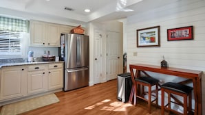 A kitchen with white cabinets, a stainless steel refrigerator, a wooden dining table with two chairs, and a small trash can.