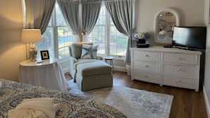 A bedroom with a large window, a television, a wicker dresser, a chair, and a patterned rug.