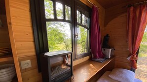 A cozy cabin room with a wooden interior, a window with curtains, a bed with white bedding, and a small heater on the windowsill. - Bohemian caravan (Chassenard)