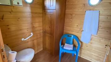Ein kleines Badezimmer in einer Holzhütte mit Toilette, einem blauen Plastikstuhl und zwei runden Fenstern.