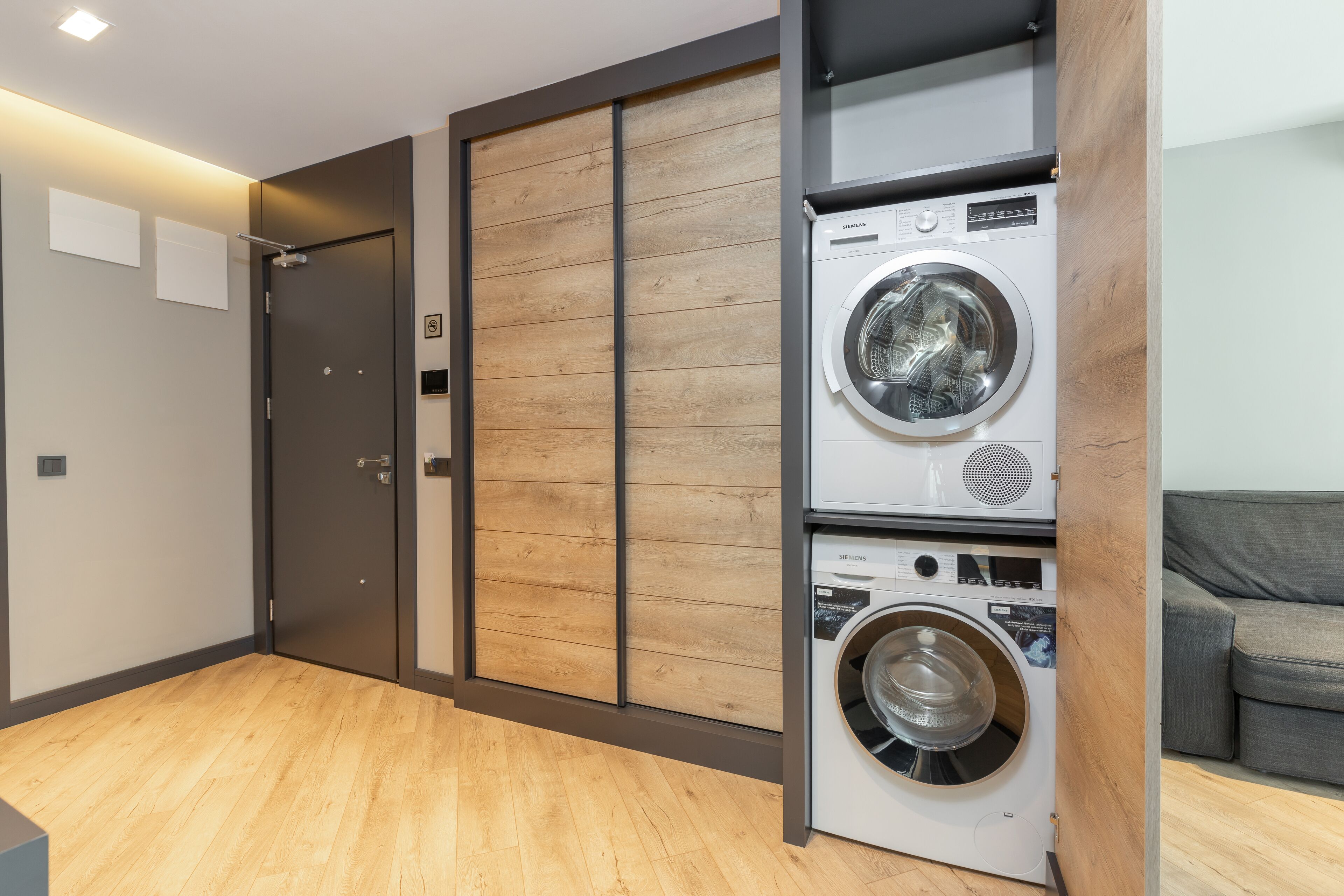 Un lavadero con dos lavadoras apiladas, un panel de madera en la pared, una puerta gris y un sofá gris al fondo.