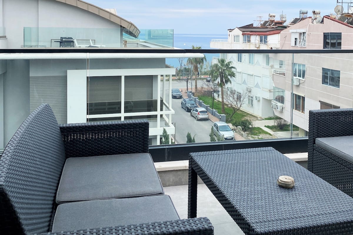 Ein Balkon mit Korbmöbeln, einer Glastrennwand und Blick auf eine Straße und Gebäude.