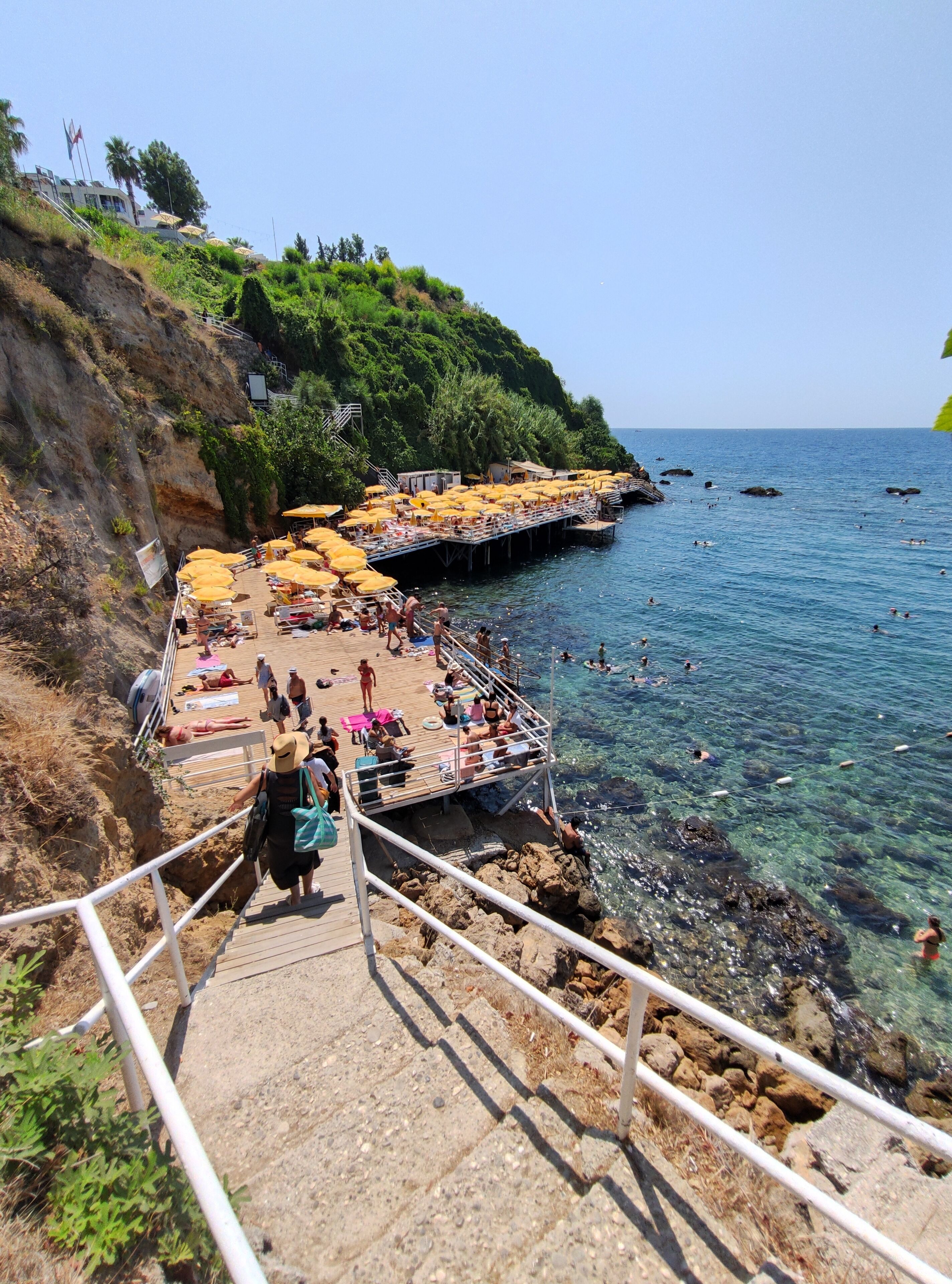 Une promenade en bord de mer qui mène à une plage où l’eau est claire, où l’on trouve des chaises longues et des parasols.