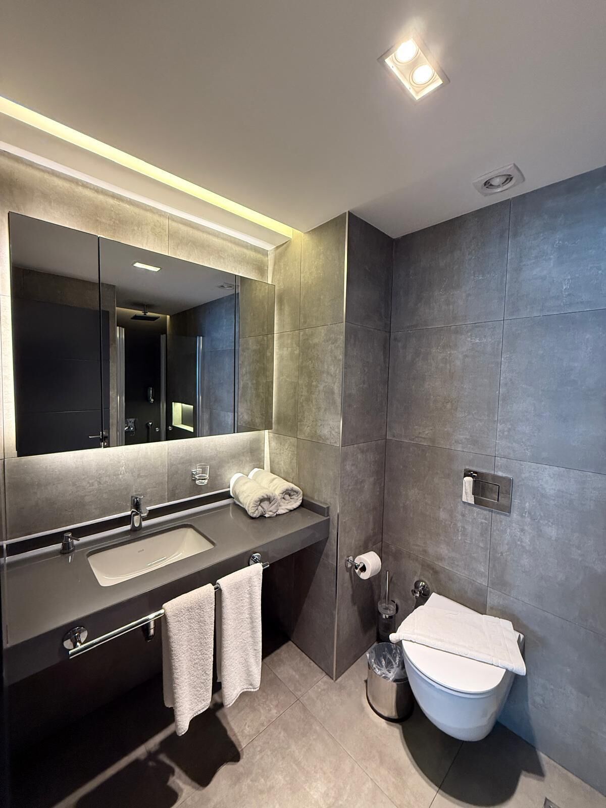 A modern bathroom with a large mirror, dark tiles, a sink, and a toilet.