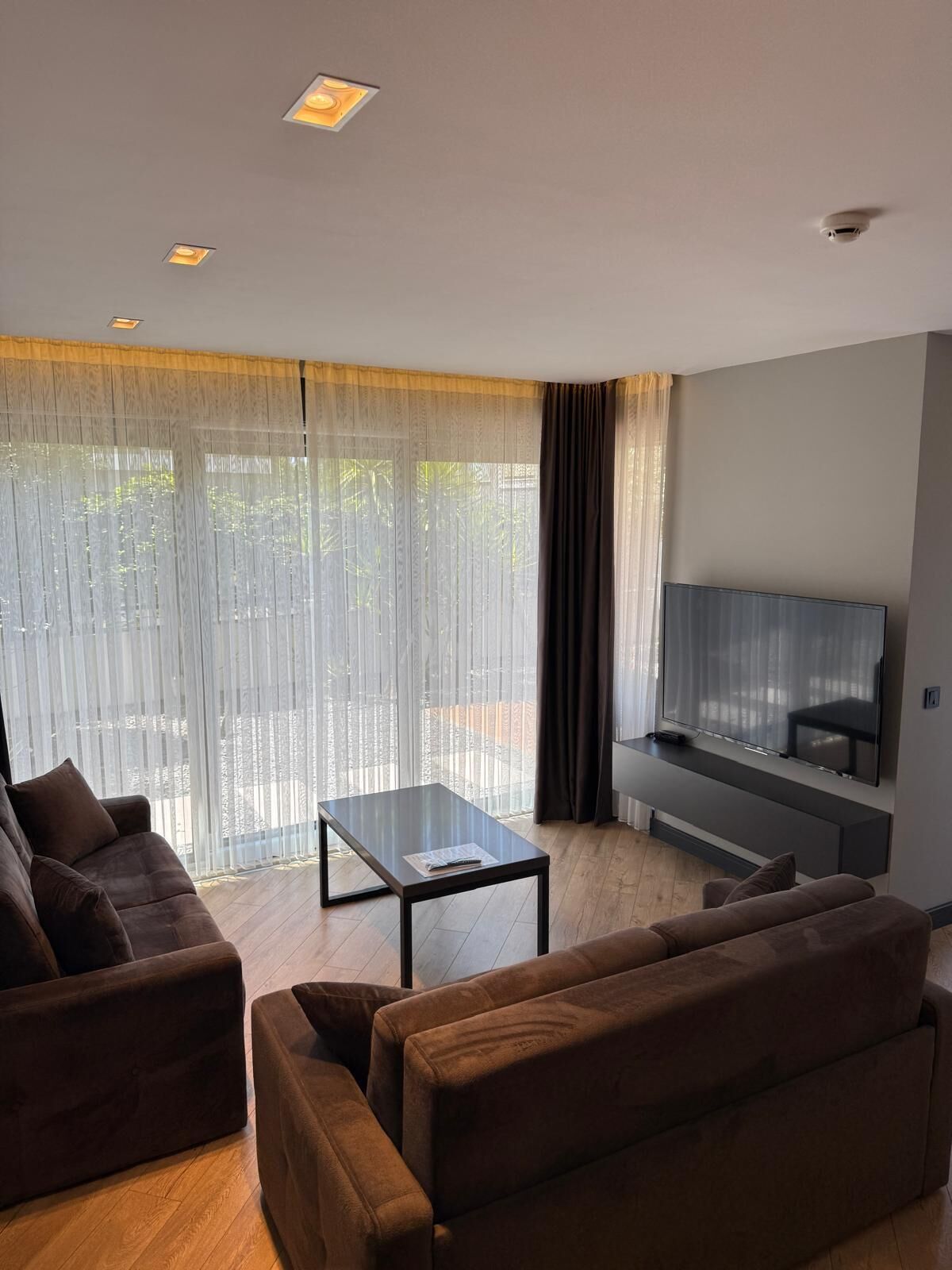 A modern living room with a flat-screen TV mounted on the wall, a glass coffee table, and two brown sofas.