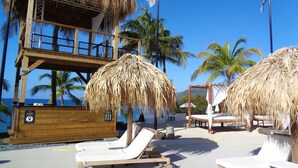 Eine Strandlounge mit reetgedeckten Sonnenschirmen, Liegestühlen und einer Holzhütte mit einem Balkon mit Meerblick.