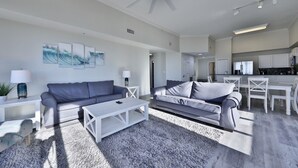Ein modernes Wohnzimmer mit zwei grauen Sofas, einem weißen Couchtisch und einem grauen Shaggy-Teppich. Es gibt einen Deckenventilator, einen Essbereich mit weißem Tisch und Stühlen sowie eine Küche mit weißen Schränken und Geräten.