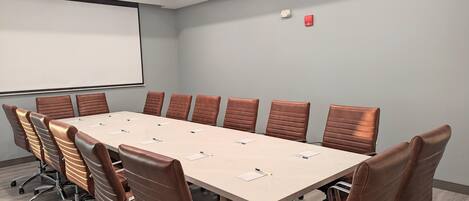 A conference room with a long table, brown leather chairs, a projector screen, and a fire alarm.
