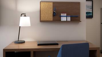 A modern office desk with a laptop, a desk lamp, a corkboard with keys and a notepad, and a framed picture on the wall.