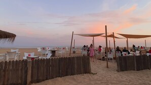 A beachside seating area with white plastic chairs and tables, wooden fences, and string lights.