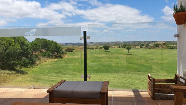 A patio with a wooden bench and a table, overlooking a green field and trees.