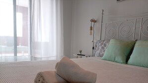 A neatly made bed with a folded towel and a 'Welcome' sign, a bedside table with a lamp, and a view through a window with sheer curtains.