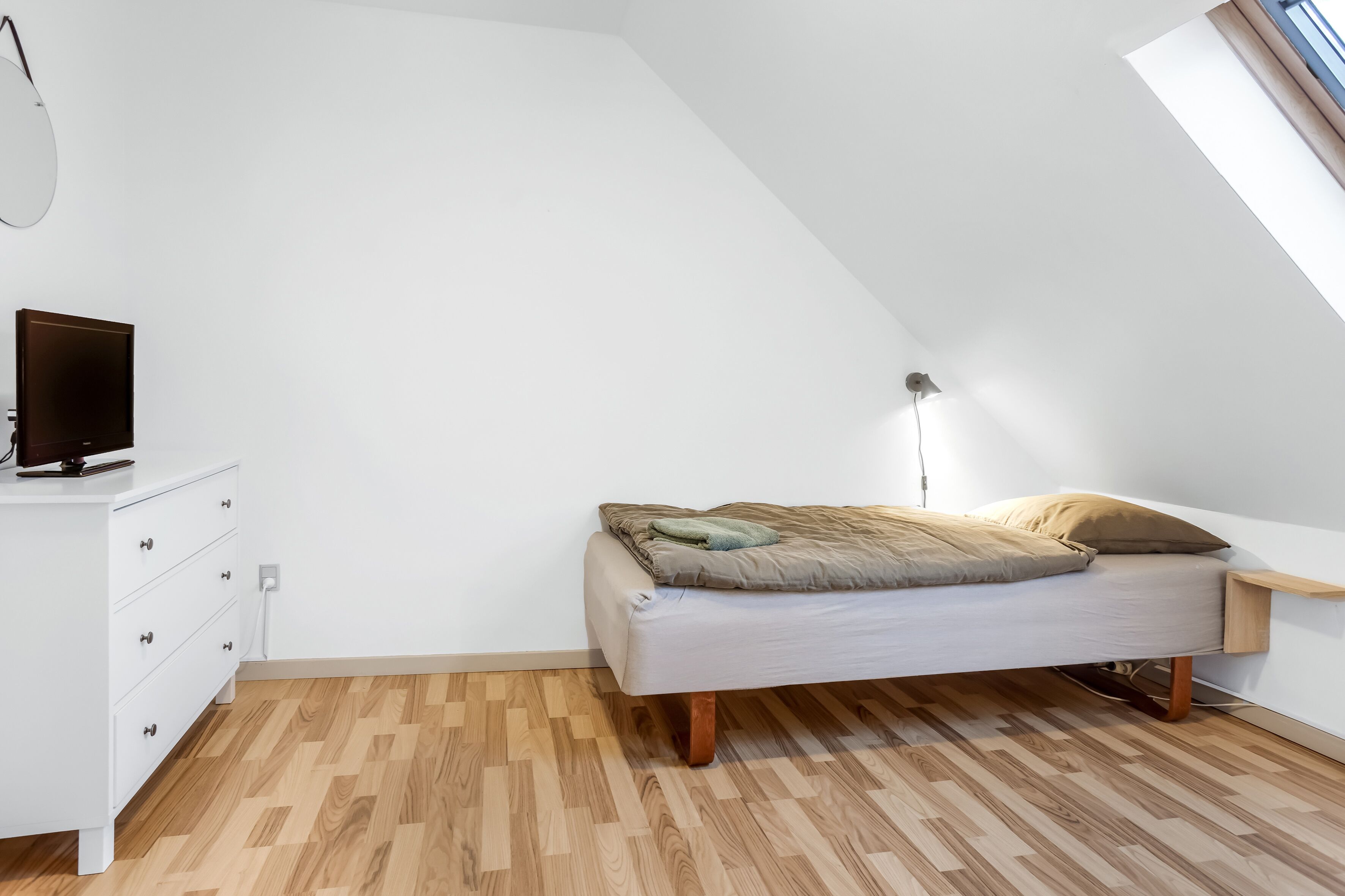A small, modern bedroom with a sloped ceiling, a bed with a light-colored mattress and pillows, a white dresser with drawers, and a small bedside table.