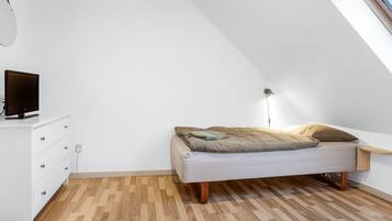 A small, modern bedroom with a sloped ceiling, a bed with a light-colored mattress and pillows, a white dresser with drawers, and a small bedside table.