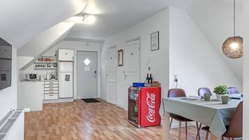 Una cuina moderna amb armaris blancs, un refrigerador i un microones. Hi ha una zona de dinar amb taula i cadires, i un refrigerador Coca-Cola.