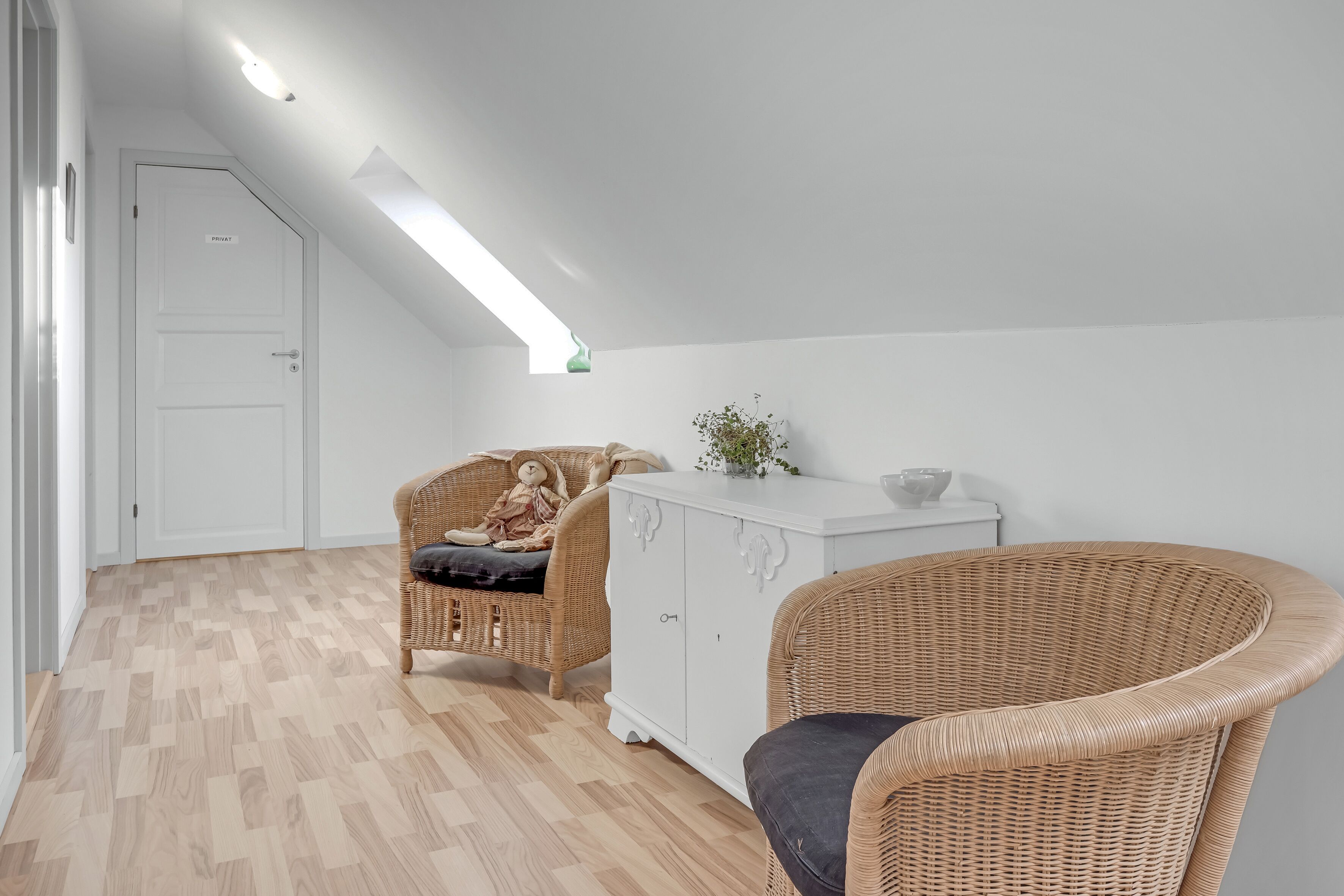 Une pièce avec un plafond en pente, une porte blanche, une chaise en osier, un meuble blanc et une petite plante.