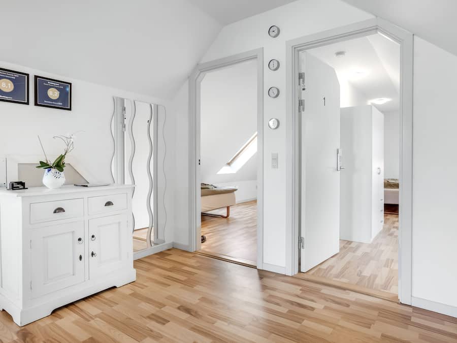 Un cuarto amplio con piso de madera, paredes blancas y un mueble blanco con cajones. Se observan dos diplomas enmarcados en la pared y una planta encima del mueble.