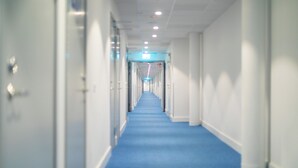 A long, narrow hallway with a blue carpet, white walls, and doors on both sides. -  ApartDirect Arlandastad (Arlandastad)