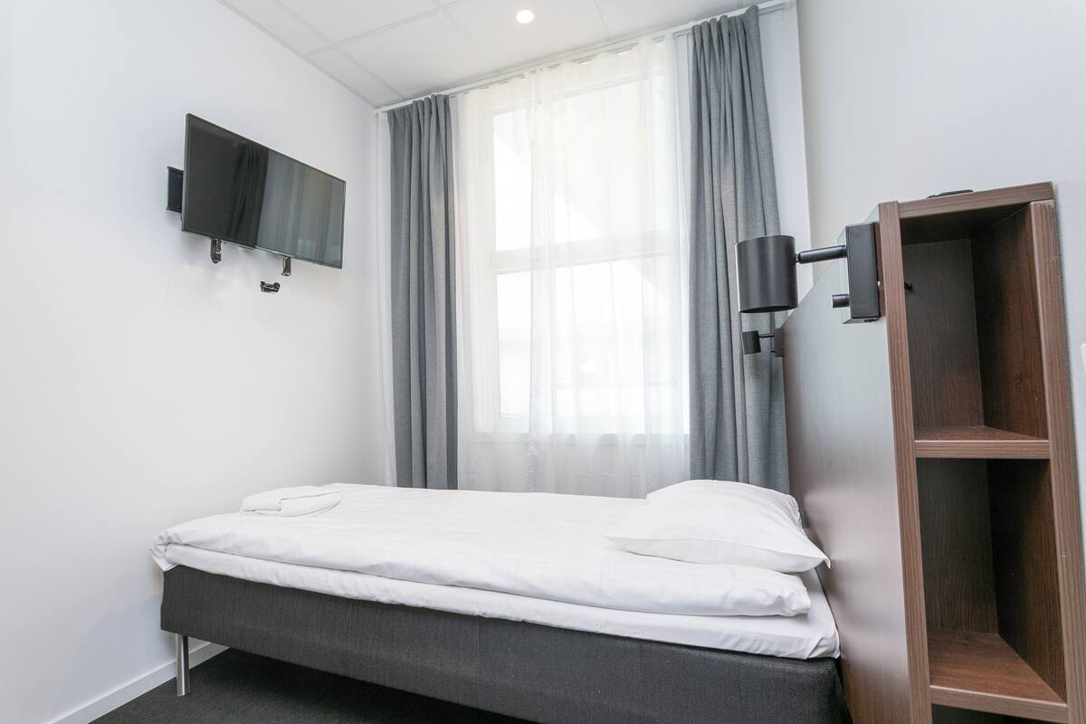 A single bed with white bedding, a mounted TV, a wooden wardrobe, and a window with sheer curtains.