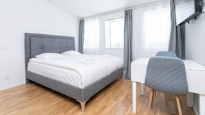 A modern bedroom with a grey bed, white bedding, a grey chair, and a white desk. -  ApartDirect Arlandastad (Arlandastad)