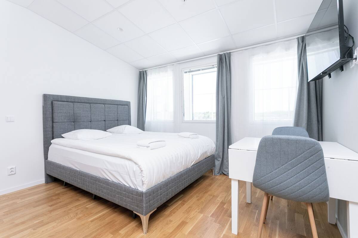 A modern bedroom with a grey bed, white bedding, a grey chair, and a white desk.