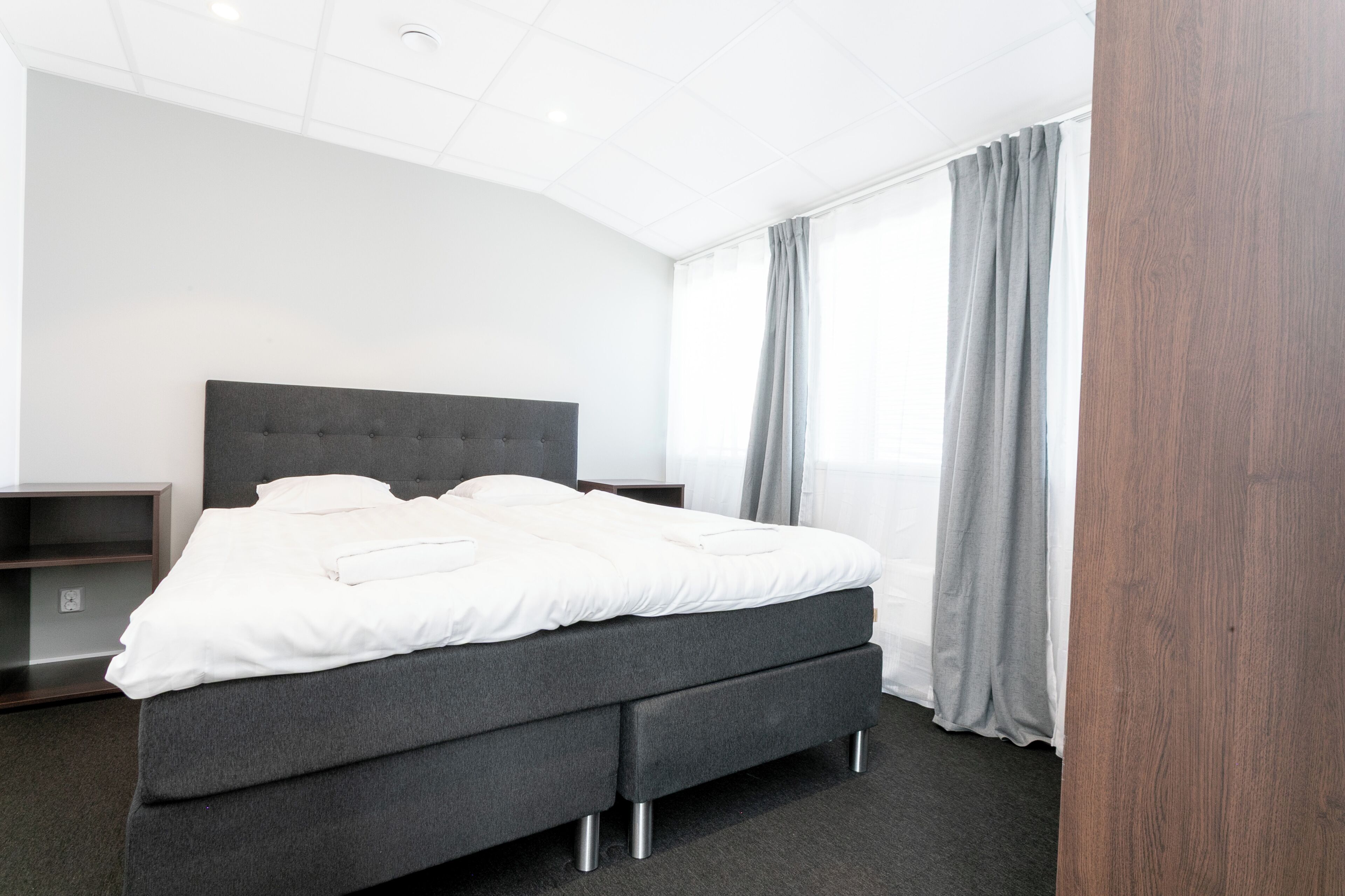 Una habitación de hotel moderna con una cama grande, ropa de cama gris, un cabecero oscuro y una cortina gris.