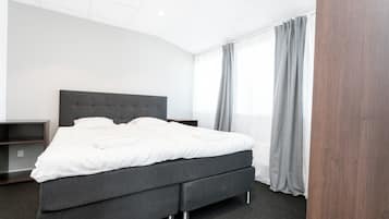 Ett modernt hotellrum med en stor säng, grå sängkläder, en mörk sänggavel och en grå gardin.