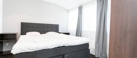 Una habitación de hotel moderna con una cama grande, ropa de cama gris, un cabecero oscuro y una cortina gris.