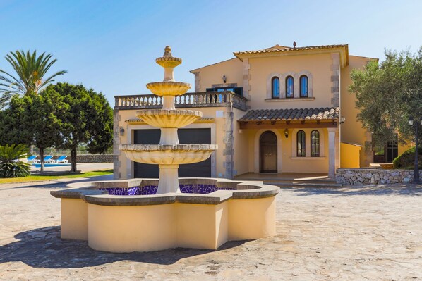 A fountain with multiple tiers in front of a building with a tiled roof and arched windows.