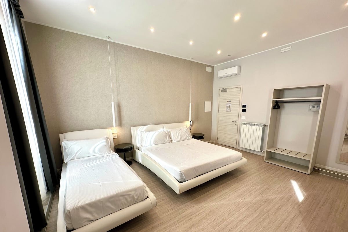 A hotel room with two beds, a wardrobe, a radiator, and a small bedside table with a lamp.