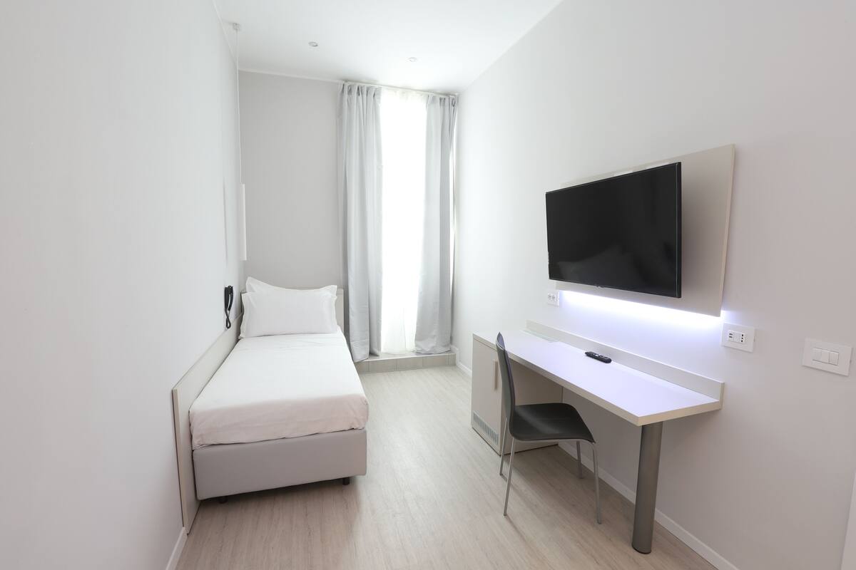 A hotel room with a bed, a desk, a chair, a TV, and a window with curtains.