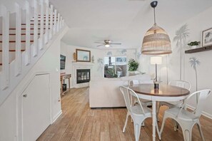 Una sala moderna con un sofá blanco, una mesa de comedor de madera y sillas blancas. Hay una chimenea, un ventilador de techo y un ventanal con vistas a palmeras.