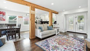 Un salón espacioso con un techo alto de vigas de madera, un sofá color beige, un tapete estampado y un comedor con mesa y sillas de madera.
