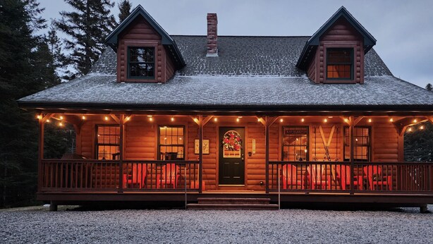 Uma cabana de toras com varanda, duas chaminés e telhado coberto de neve.