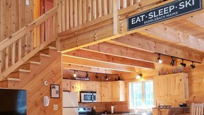 Interior rústico de uma cabana com escada de madeira, cozinha com armários de madeira, área de refeições com mesa e cadeiras, e um letreiro com a inscrição "EAT, SLEEP, SKI".