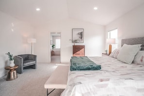 Une chambre moderne avec un grand lit, une table de chevet, une chaise et un bureau. Une fenêtre laisse entrer la lumière naturelle et une porte donne accès à une autre pièce.