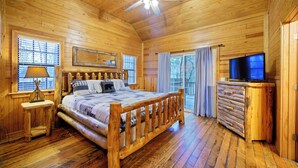 A wooden cabin bedroom with a bed, a ceiling fan, a TV, and a window with blinds.