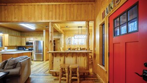 A wooden cabin interior with a kitchen, dining area, and a red door.