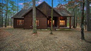 A rustic log cabin with a front porch, surrounded by tall trees.