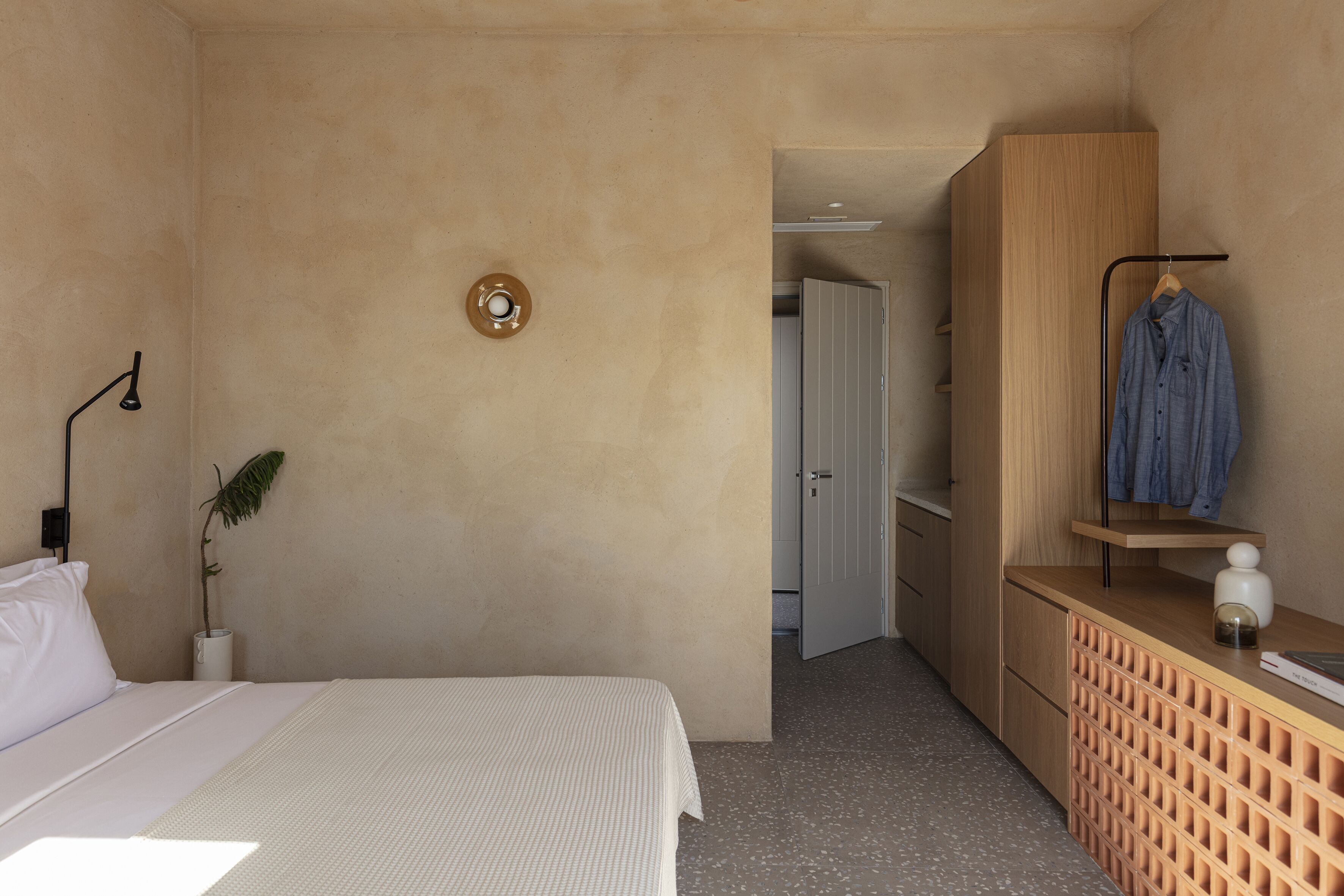A bedroom with a bed, a bedside lamp, a small plant, a wooden wardrobe, and a hanging rack with clothes.