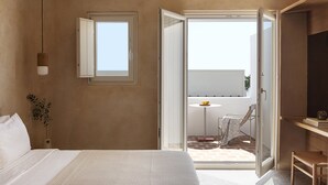 A bedroom with a bed, a bedside table, a hanging light fixture, a window, and an open door leading to a balcony with a table and chairs. - Ethos Vegan Suites (Santorini)