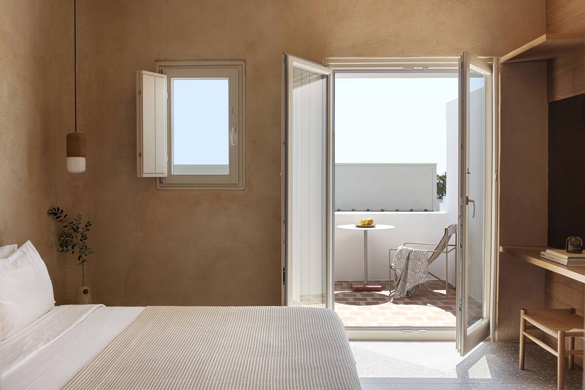 Ein Schlafzimmer mit einem Bett, einem Nachttisch, einer Hängeleuchte, einem Fenster und einer offenen Tür, die auf einen Balkon mit Tisch und Stühlen führt.