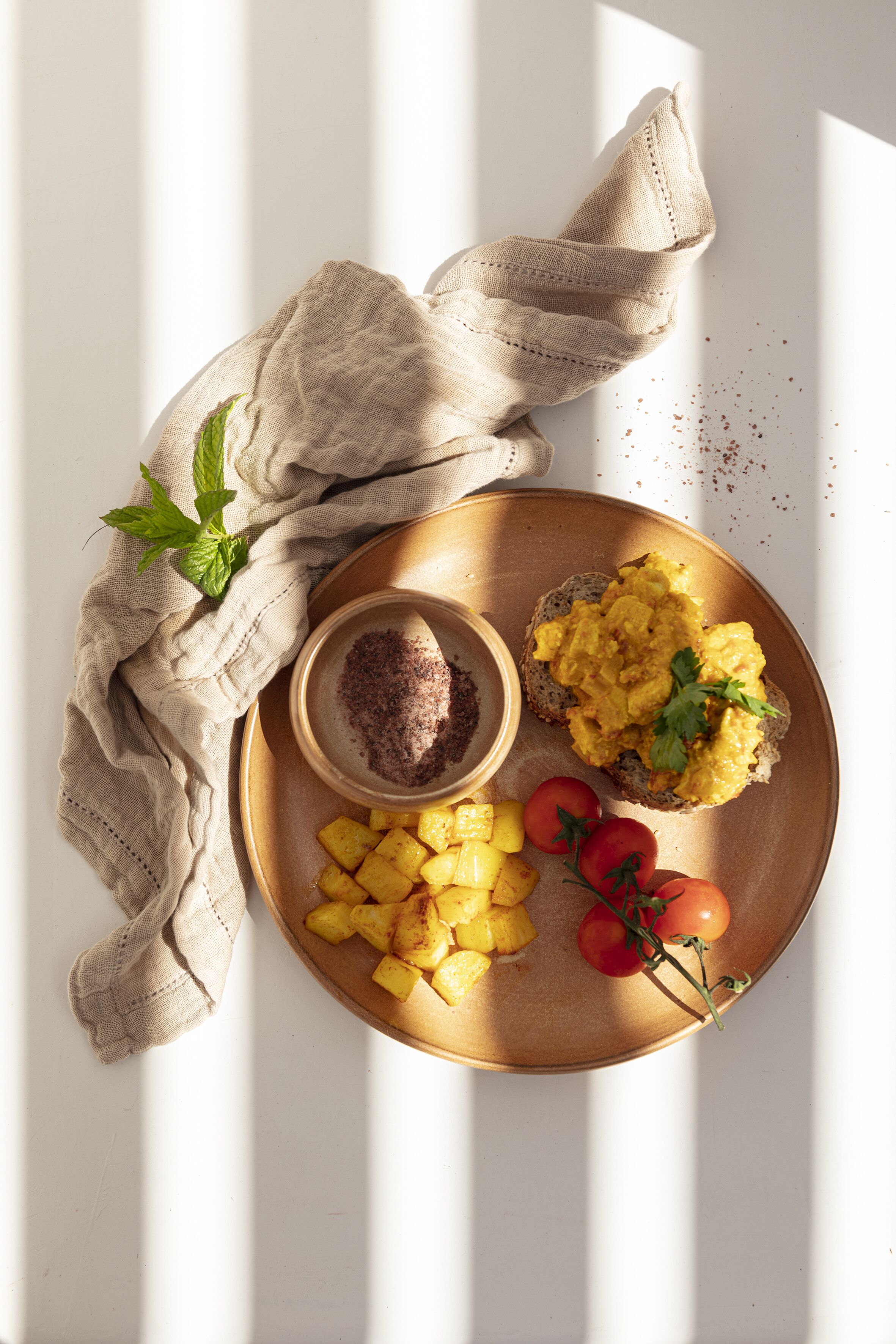 A breakfast plate with scrambled eggs, pineapple chunks, and cherry tomatoes on a wooden board, accompanied by a bowl of chocolate spread and a cloth napkin.
