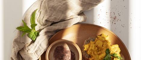 A breakfast plate with scrambled eggs, pineapple chunks, and cherry tomatoes on a wooden board, accompanied by a bowl of chocolate spread and a cloth napkin.