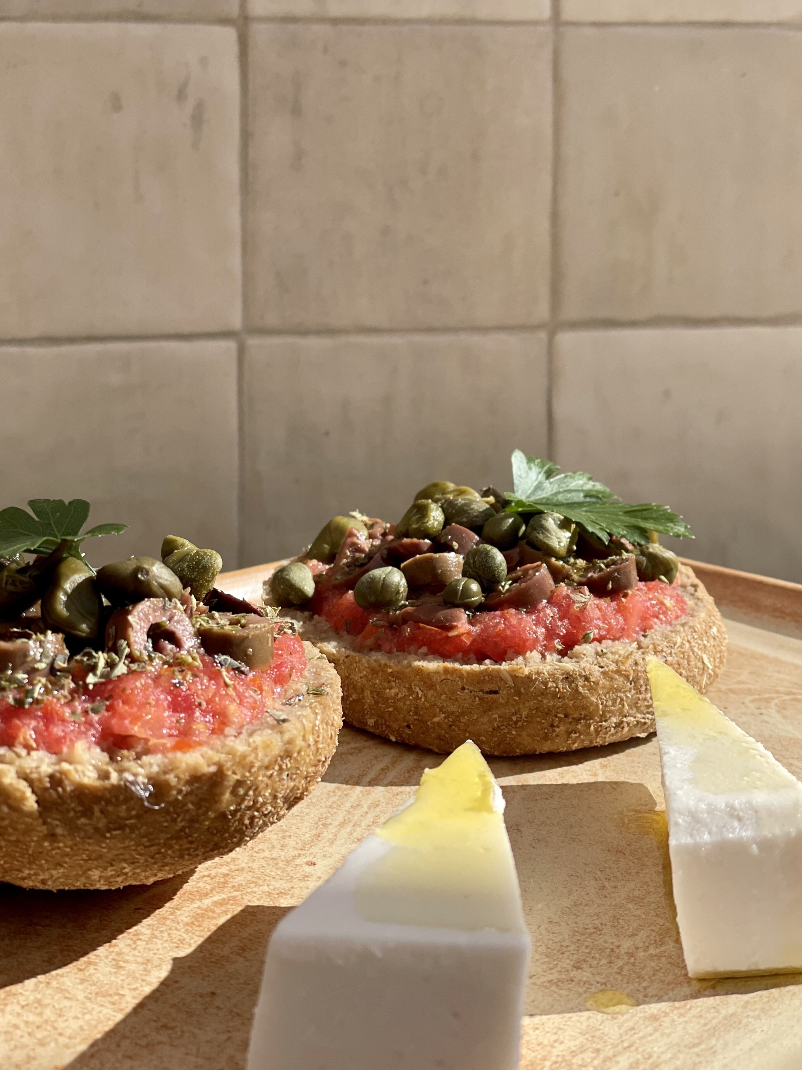 Two open-faced sandwiches on a wooden board, garnished with herbs and capers, with a knife and a wedge of cheese on the side.